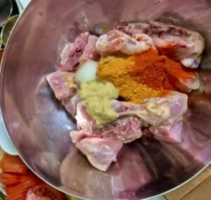 Raw chicken pieces in a steel bowl with turmeric, red chili powder, salt, and ginger garlic paste for marination.